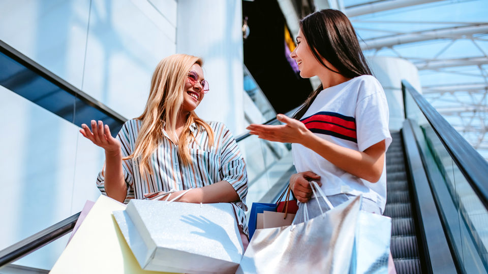 2 female shoppers carrying shopping bags riding an escalator in a retail shopping mall