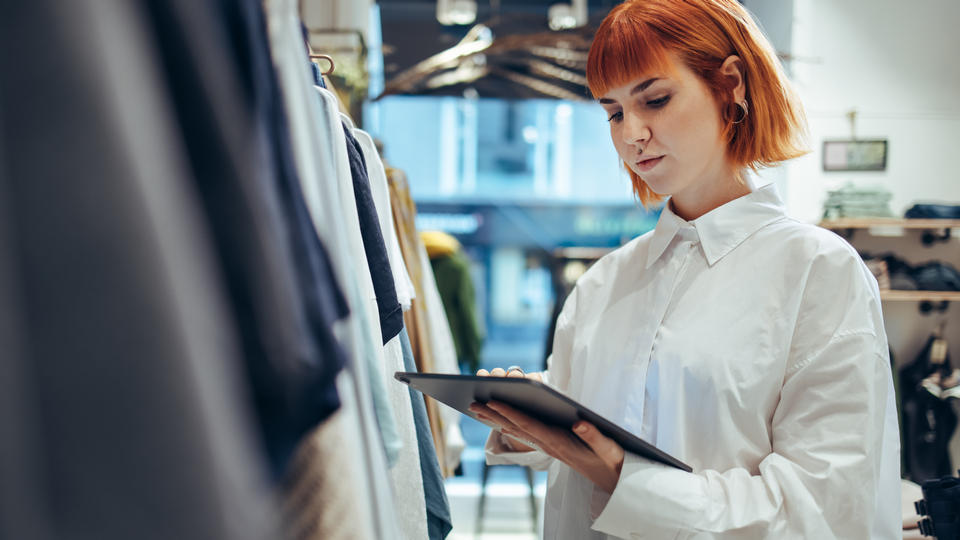 young woman with piercings and red hair wearing white shirt using tablet to take inventory in retail clothing store